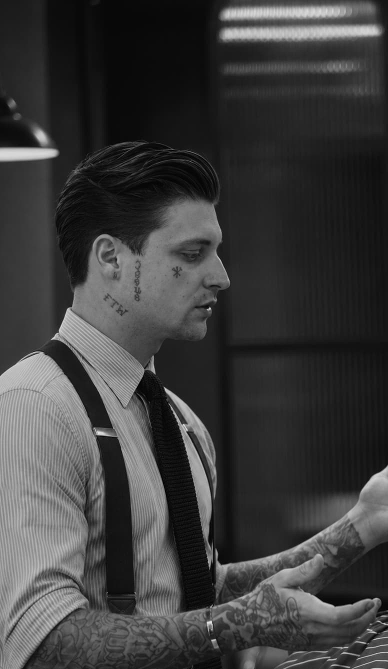 Retrato em preto e branco do fundador de perfil na barbearia, de camisa listrada, suspensórios e gravata de tricô